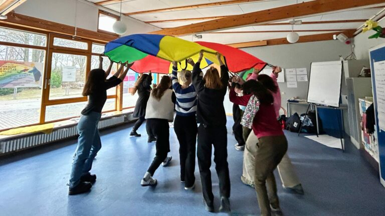 Erwachsene stehen im Kreis und werfen zusammen ein Fallschirmtuch in die Luft. Einige laufen unter dem Fallschirmtuch und durch den Kreis.