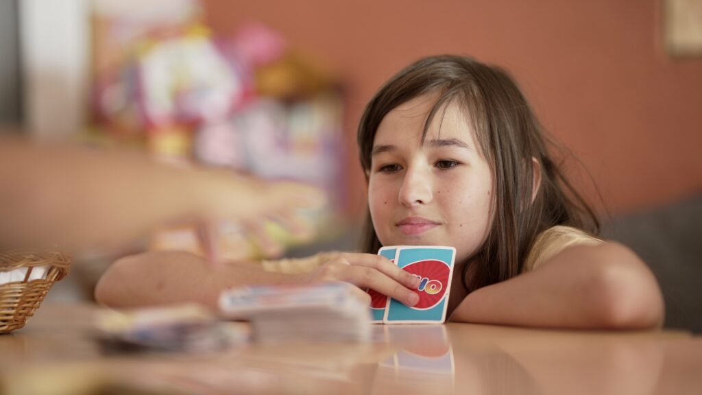 Ein Mädchen sitzt an einem Tisch und hält zwei UNO Spielkarten in der Hand.