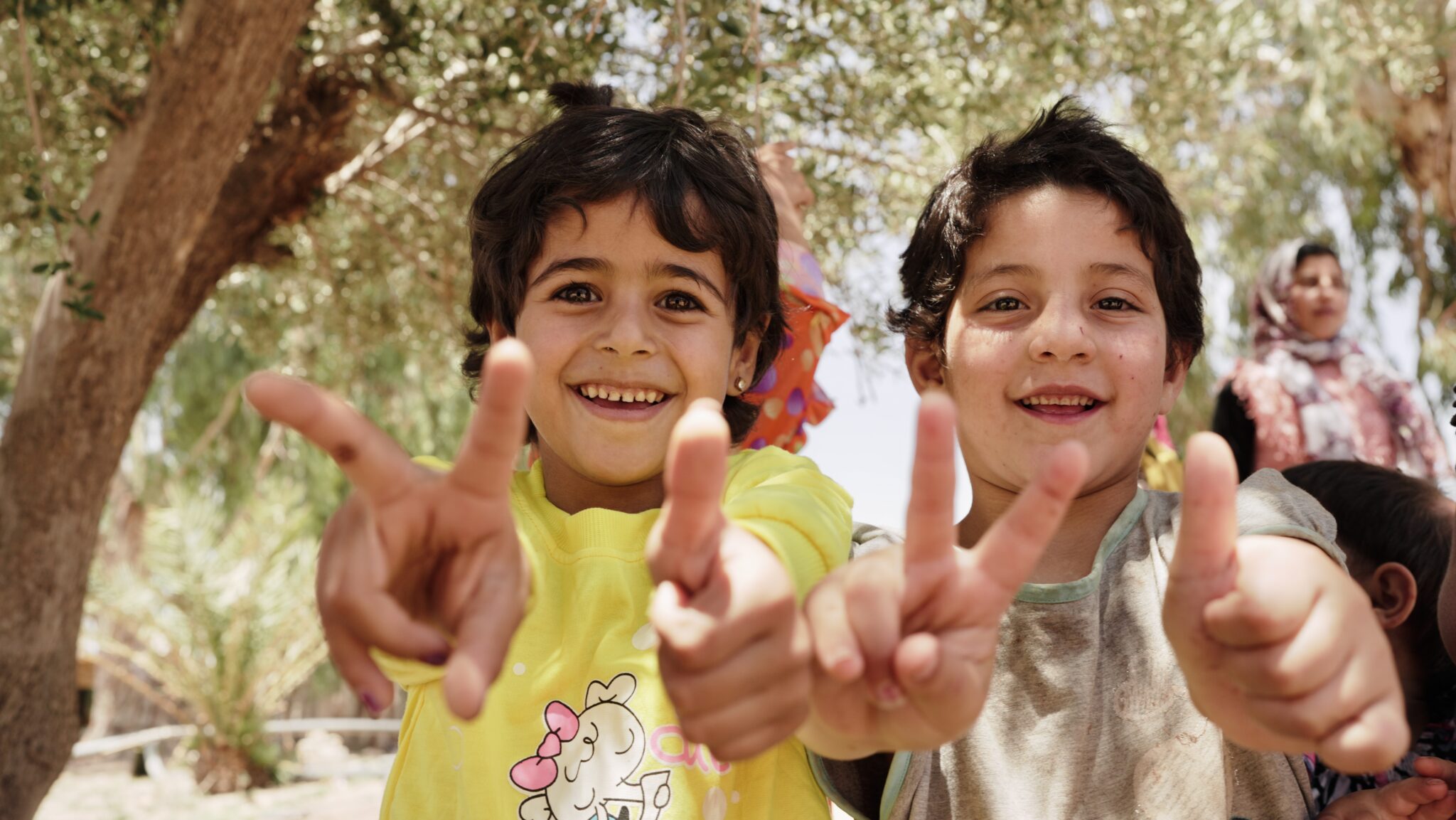 Zwei Kinder schauen in die Kamera und zeigen das Peace Zeichen und einen Daumen hoch.
