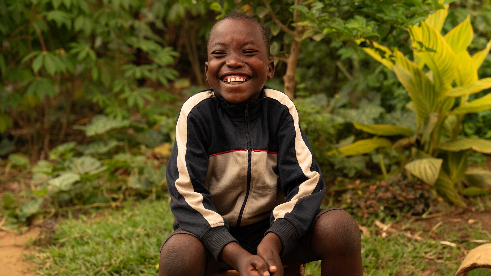 Ein Junge sitzt vor einem Busch, lächelt und zieht die Schultern hoch.
