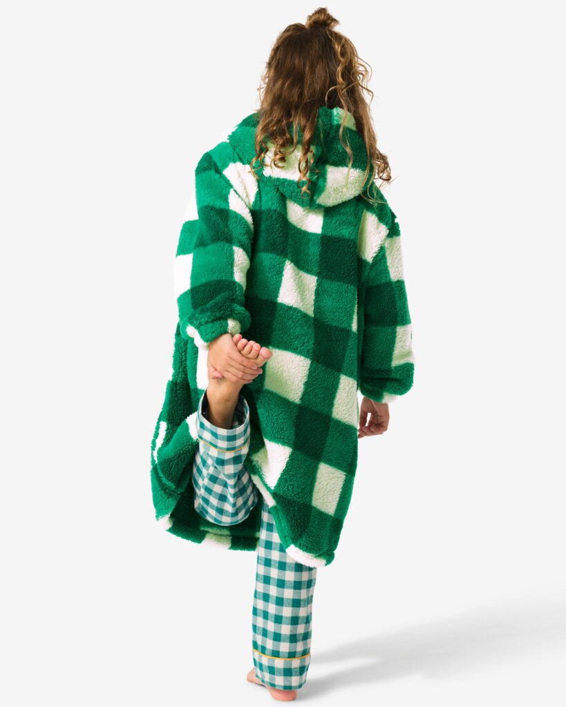 Ein Kind steht mit dem Rücken zum Betrachter und hebt ein Bein an. Es trägt einen grün-weiß karierten Pyjama und einen grün-weiß karierten Kuschelhoodie