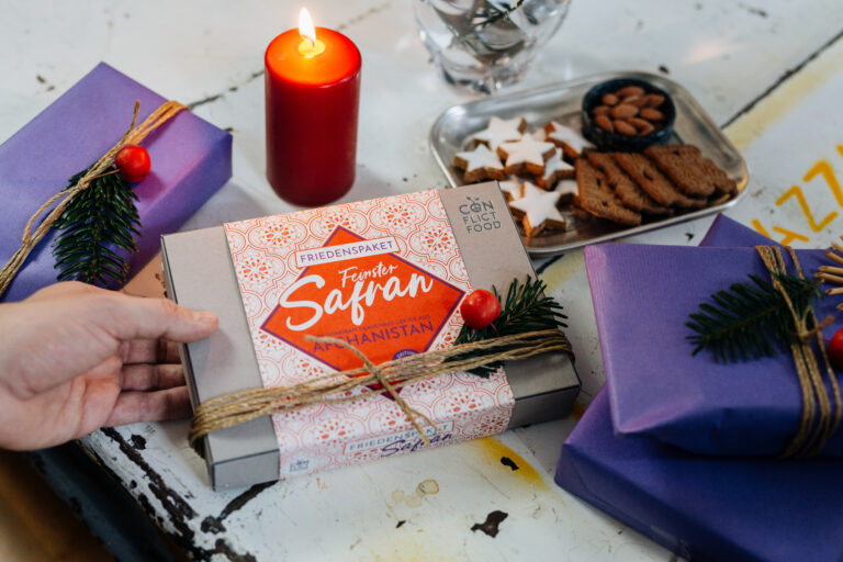 Spekulatius, Zimtsterne, Kerze, lila Geschenkpapier. In der Mitte liegt ein Friedenspaket von Conflictfood. Darin ist Safran.