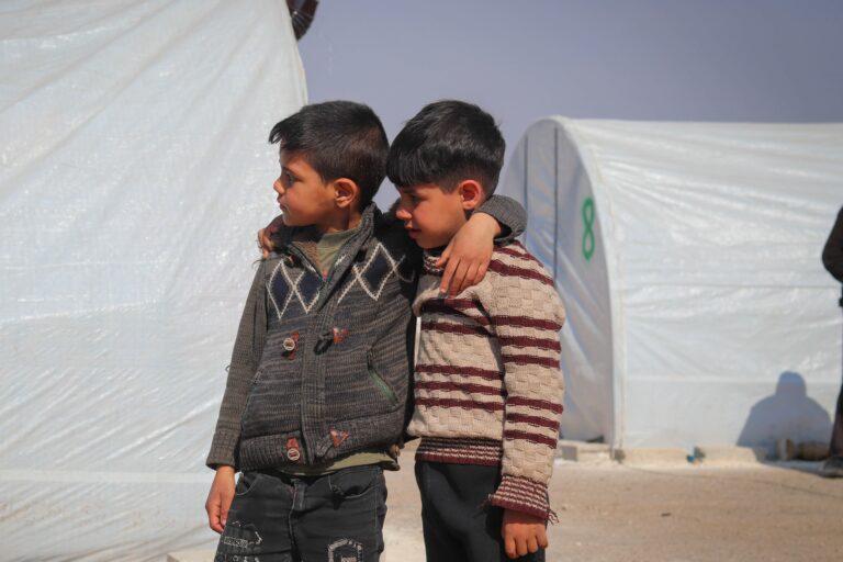 Zwei Kinder stehen vor großen weißen Zelten und umarmen sich.