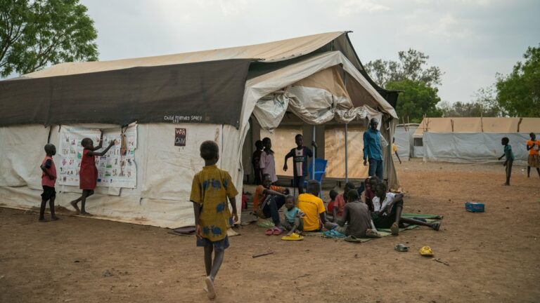 Kinder versammeln sich vor und in einem großen Zelt mit der Aufschrift „Child Friendly Space“, einige sitzen auf Matten, andere stehen daneben; im Hintergrund weitere Zelte auf staubigem Gelände.
