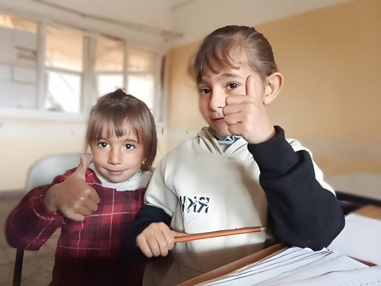 Zwei Kinder sitzen im Klassenzimmer am Tisch, halten den Daumen hoch und haben ein Heft und einen Stift vor sich.