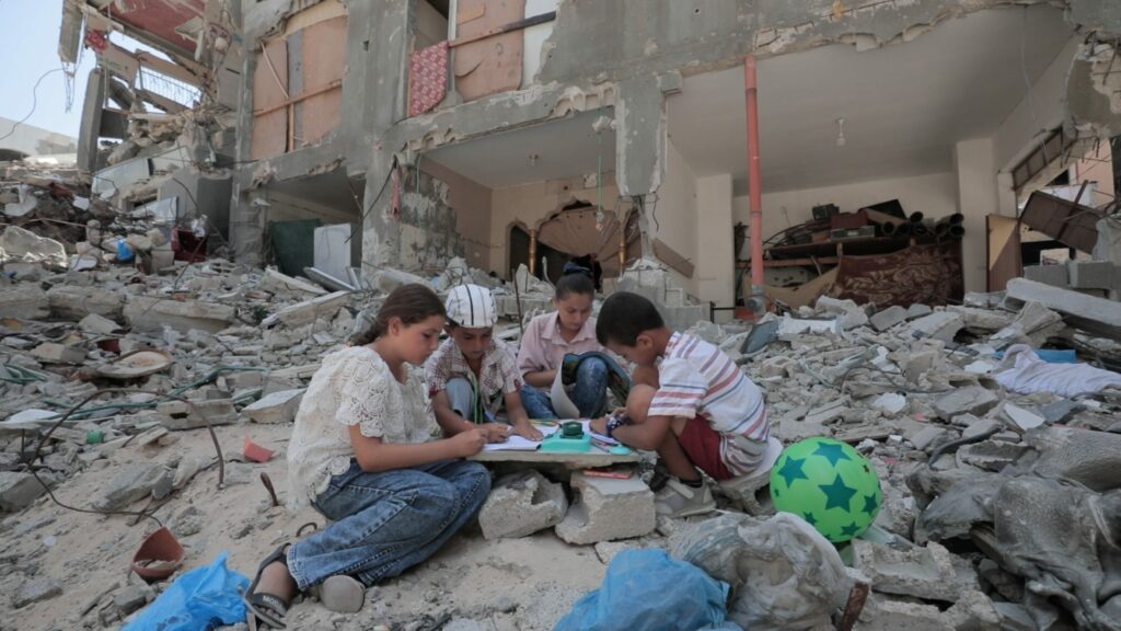Vier Kinder sitzen zwischen den Trümmern eines zerstörten Gebäudes in Gaza und malen bzw. spielen gemeinsam auf dem Boden.