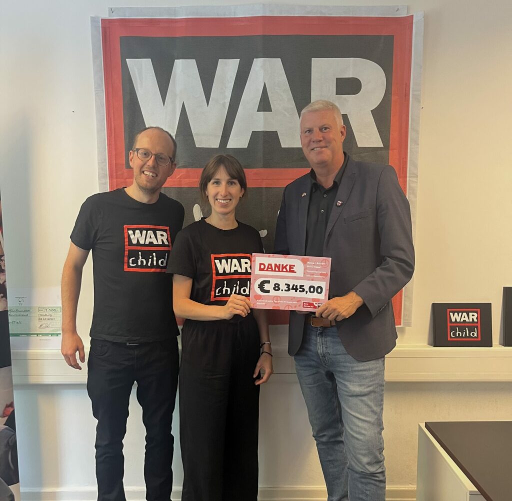 Drei Leute stehen vor einer Wand mit einer War Child Flagge und halten einen großen Scheck hoch.