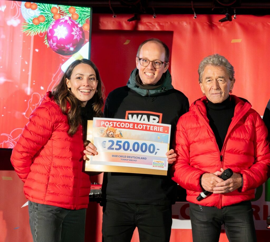 Vor einem roten Hintegrund steht ein Mann mit einem Scheck von 250.000 Euro. Links von ihm steht eine lächelnde Frau und rechts von ihm ein lächelnder älterer Mann.