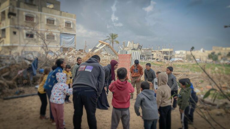 Eine Gruppe von Kindern steht gemeinsam mit zwei Erwachsenen, die einen War-Child-Pullover tragen, im Kreis. Um sie herum befinden sich Trümmer von Gebäuden.