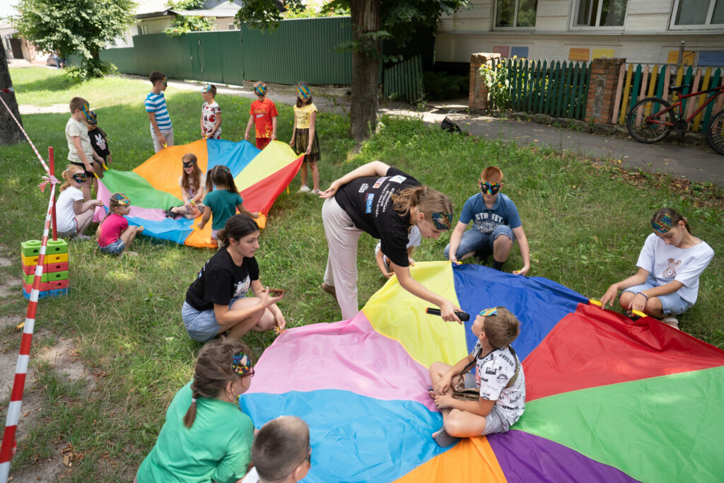 Auf einer Grünen Wiese sind zwei kleine Kindergruppen, die jeweils ein aufgespanntes buntes Tuch auf dem Boden liegen haben. Drum herum sitzen Kinder. In der Mitte der Nebeneinander gespannten Tücher sitzt jeweils ein Kind. Bei der rechten Gruppe hält eine Frau dem Kind in der Mitte ein Mikrofon zu.