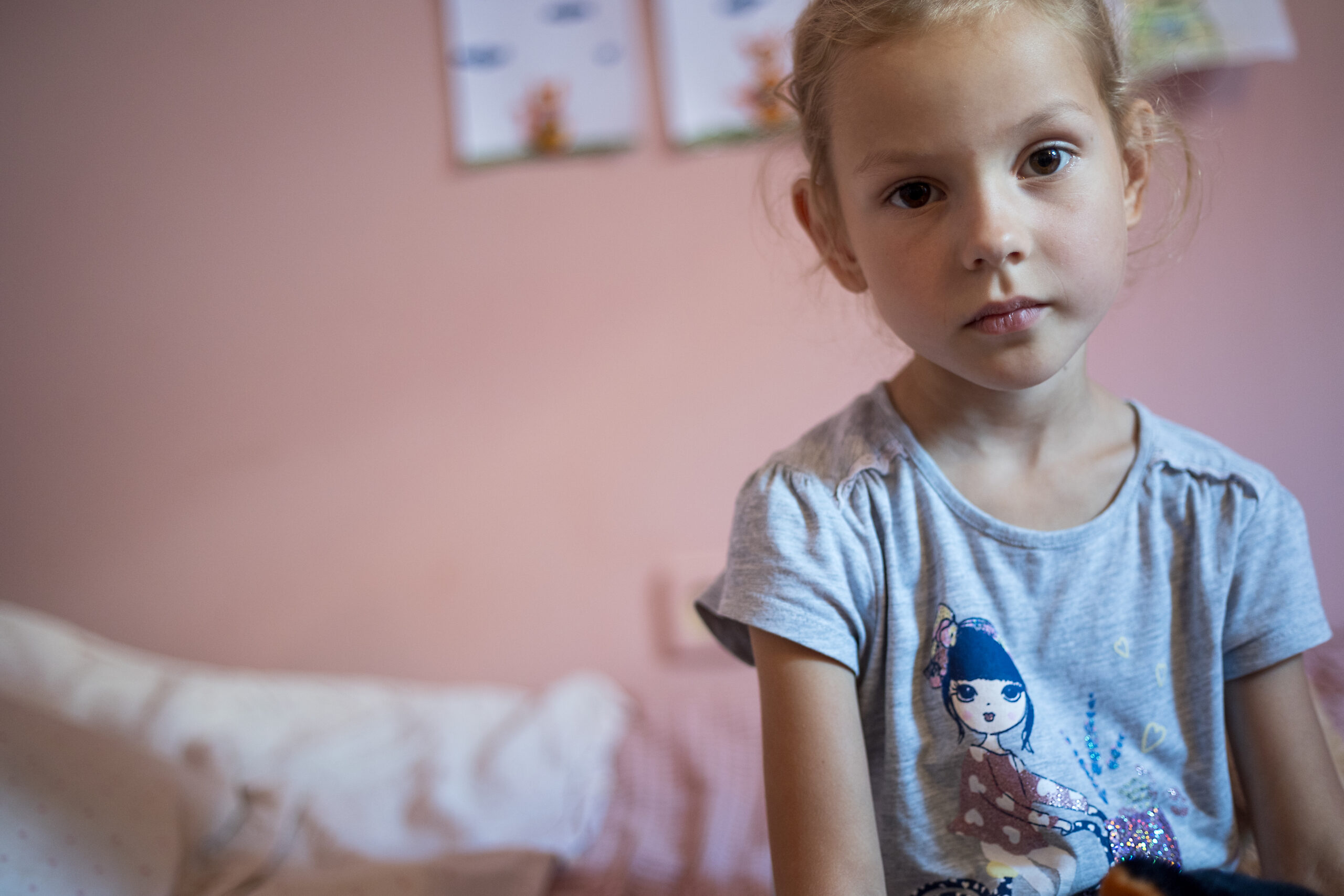 Blondes Mädchen im Alter von etwa 5 Jahren schaut trostlos. Sie sitzt in einem Rosa gestrichenen Zimmer auf ihrem Bett.