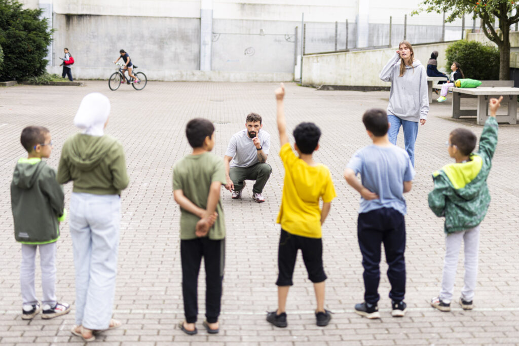 Fünf Kinder stehen auf einem gepflasterten Platz in einer Reihe. Zwei haben den Arm gehoben und melden sich. Ihnen gegenüber hockt ein Mann auf dem Boden. Neben ihm steht eine Frau.